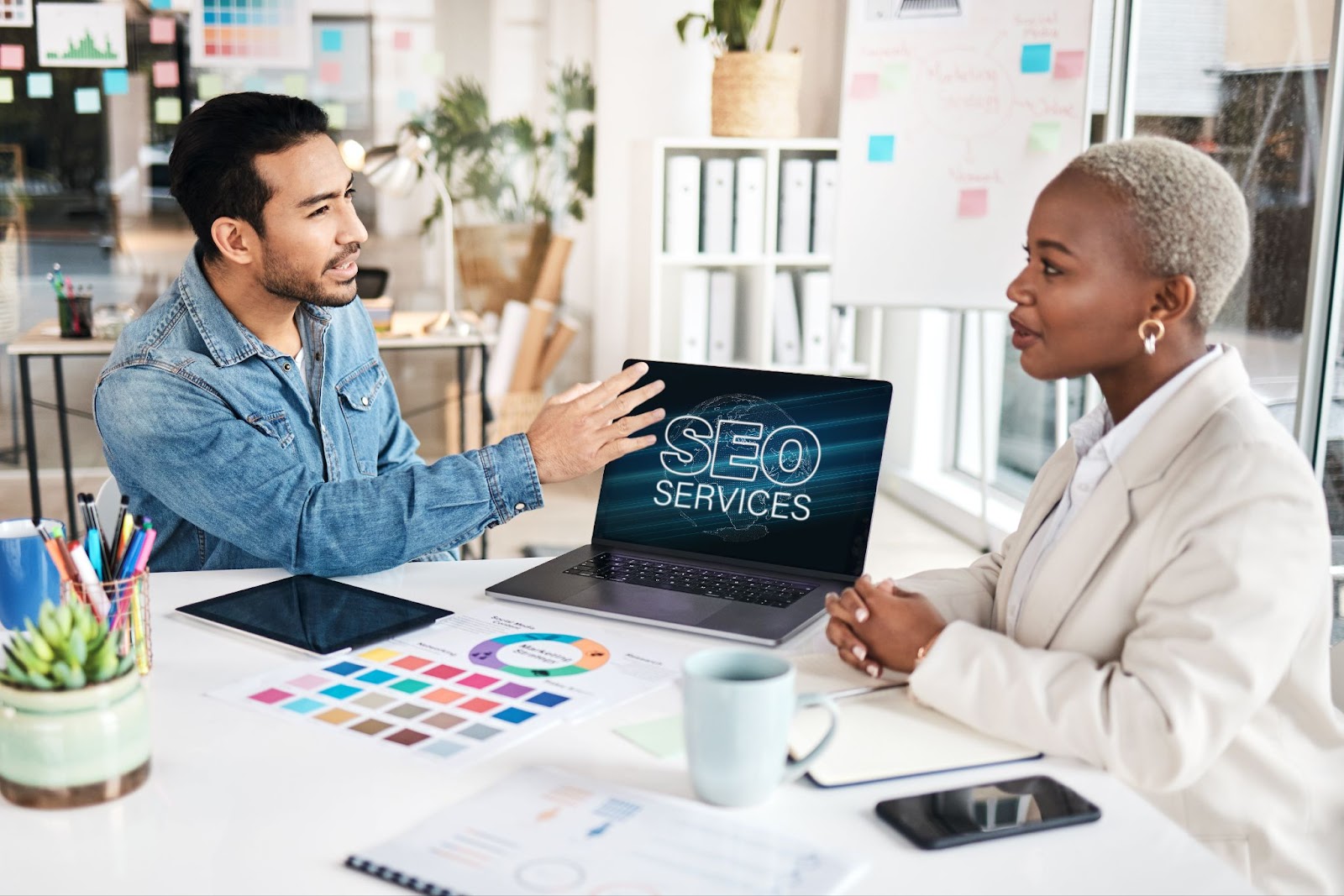 A man wearing a jean jacket touching a laptop that says “SEO services” while talking to a woman wearing white in an office.