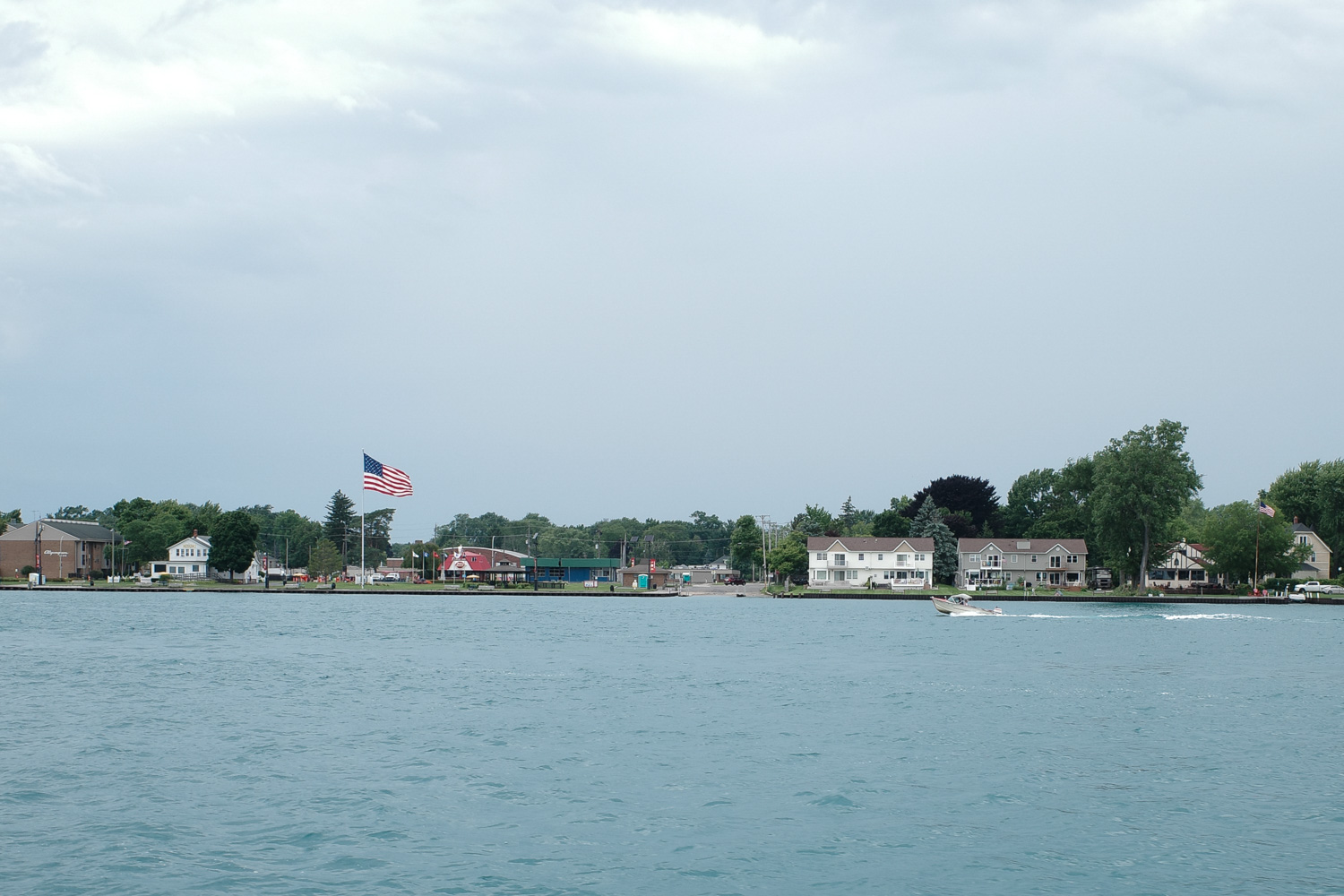 View of Algonac Michigan from ferry, with lake houses, boat speeding past, dairy queen, and tall American flag.