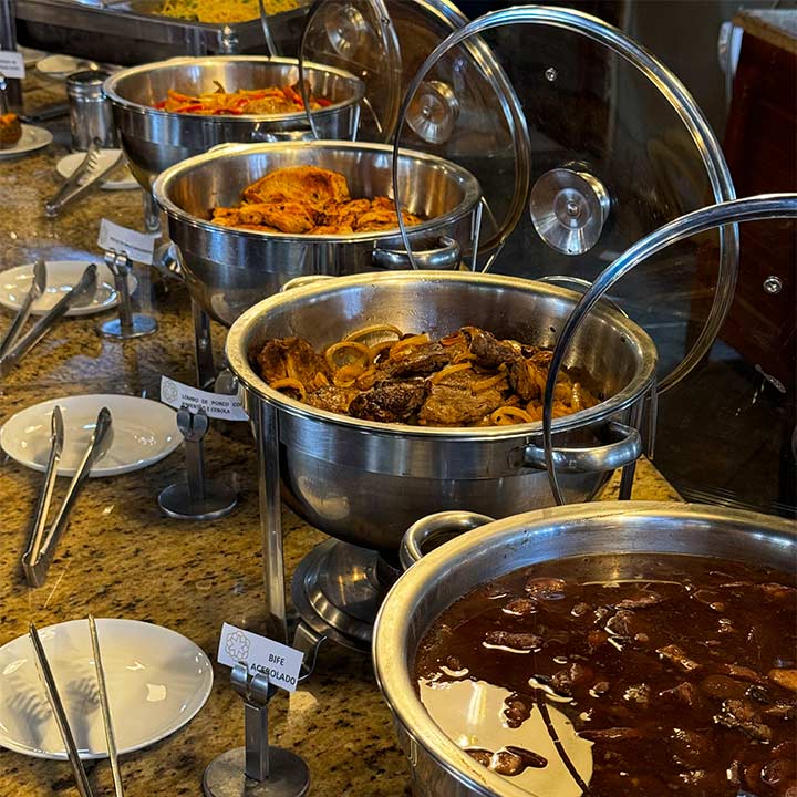 Mesa de buffet com diferentes pratos quentes, incluindo carne, massas e acompanhamentos, ideal para amantes da boa gastronomia.