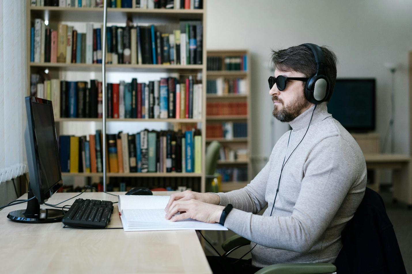 A person with visual impairment reading Braille text on a wooden desk, with a laptop, sunglasses, and an abacus nearby, indicating an accessible learning environment.