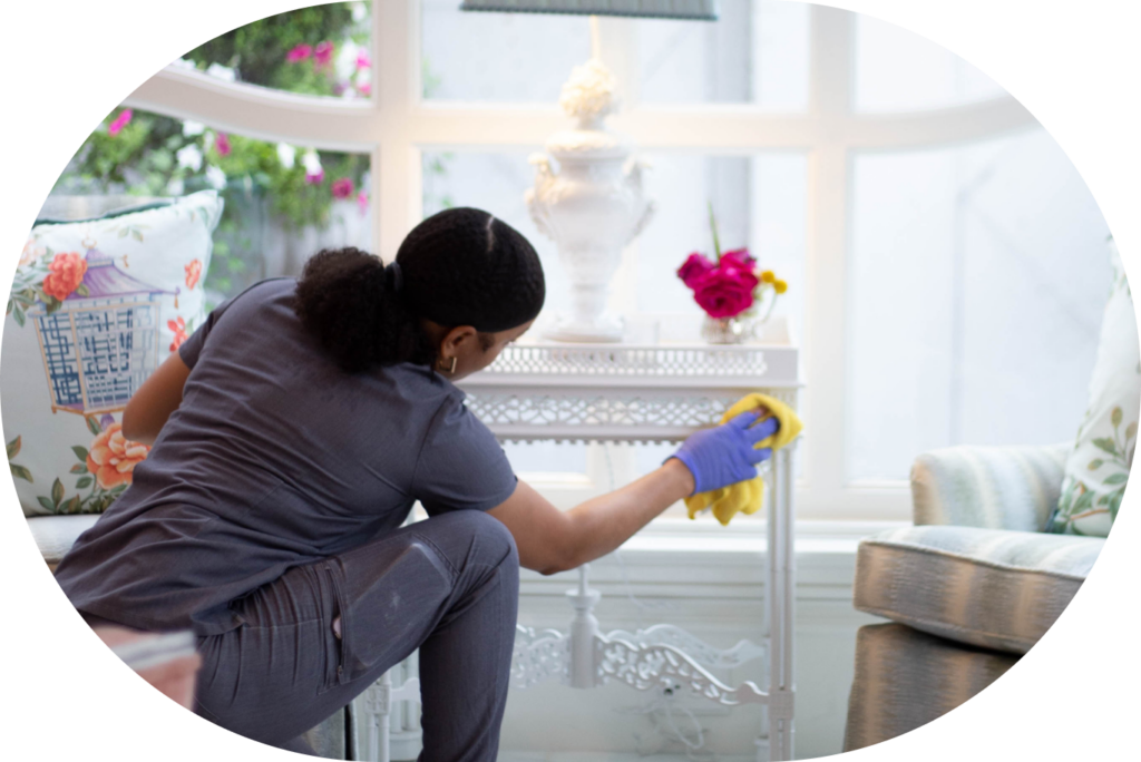 The image shows a cleaning professional wearing a grey uniform and purple gloves, carefully wiping down a decorative white side table in a bright, well-decorated living room. The cleaner is using a yellow cloth, focused on removing dust or dirt from the surface. The room is stylishly furnished with a floral-patterned pillow, a cozy chair, and a vase with fresh flowers on the table, which adds a welcoming feel. This scene represents professional cleaning services in Austin or house cleaning services in Dallas, highlighting attention to detail in maintaining a clean and organized home environment.