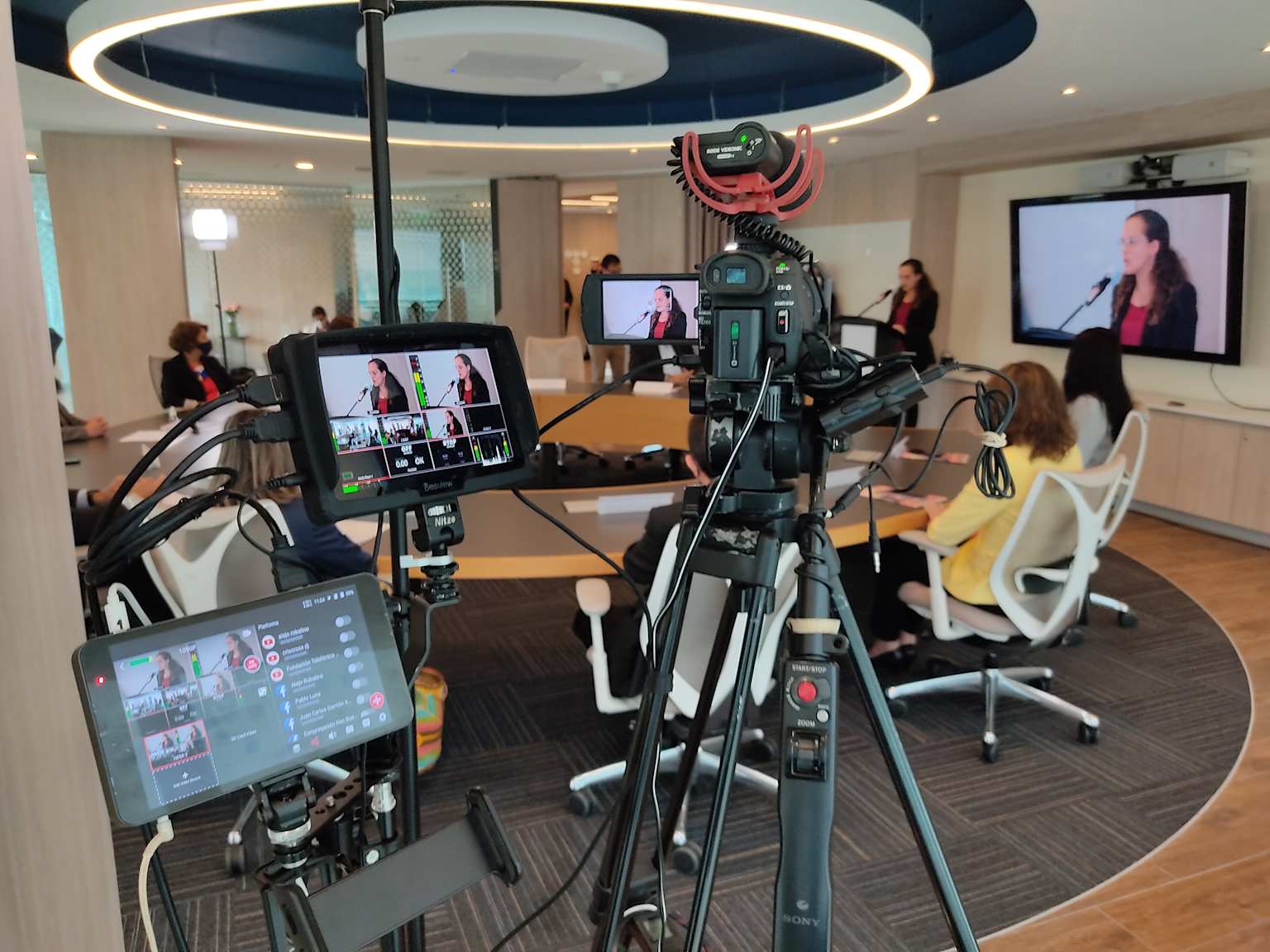 Professional corporate video production setup capturing a speaker in a modern conference room with multiple monitors and a high-quality camera