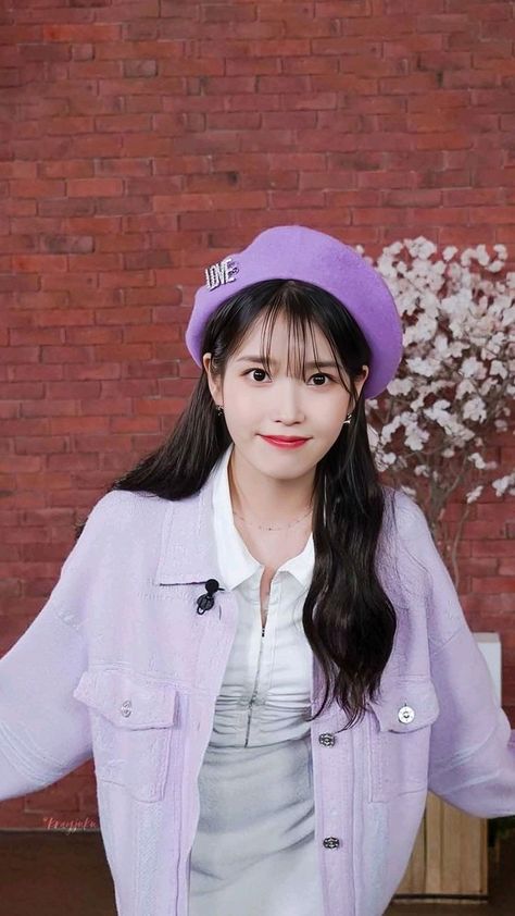 This contains an image of IU wearing a purple hat and jacket standing in front of a brick wall