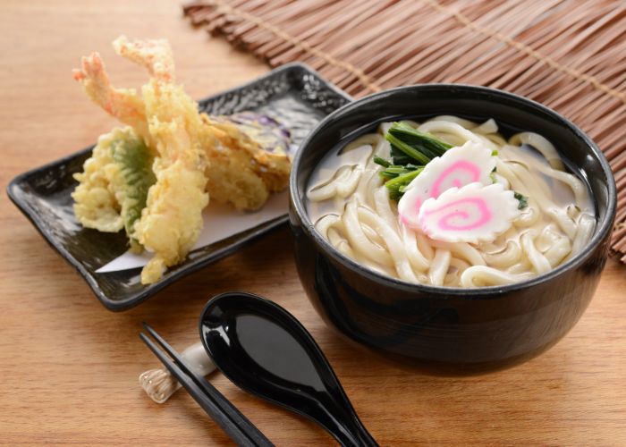 A bowl of udon topped with naruto next to a plate of assorted tempura