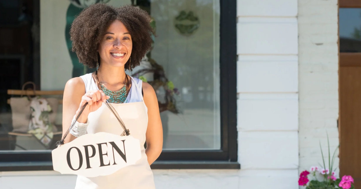 (A smiling shop owner stands outside her storefront holding an "Open" sign, ready to welcome customers.)