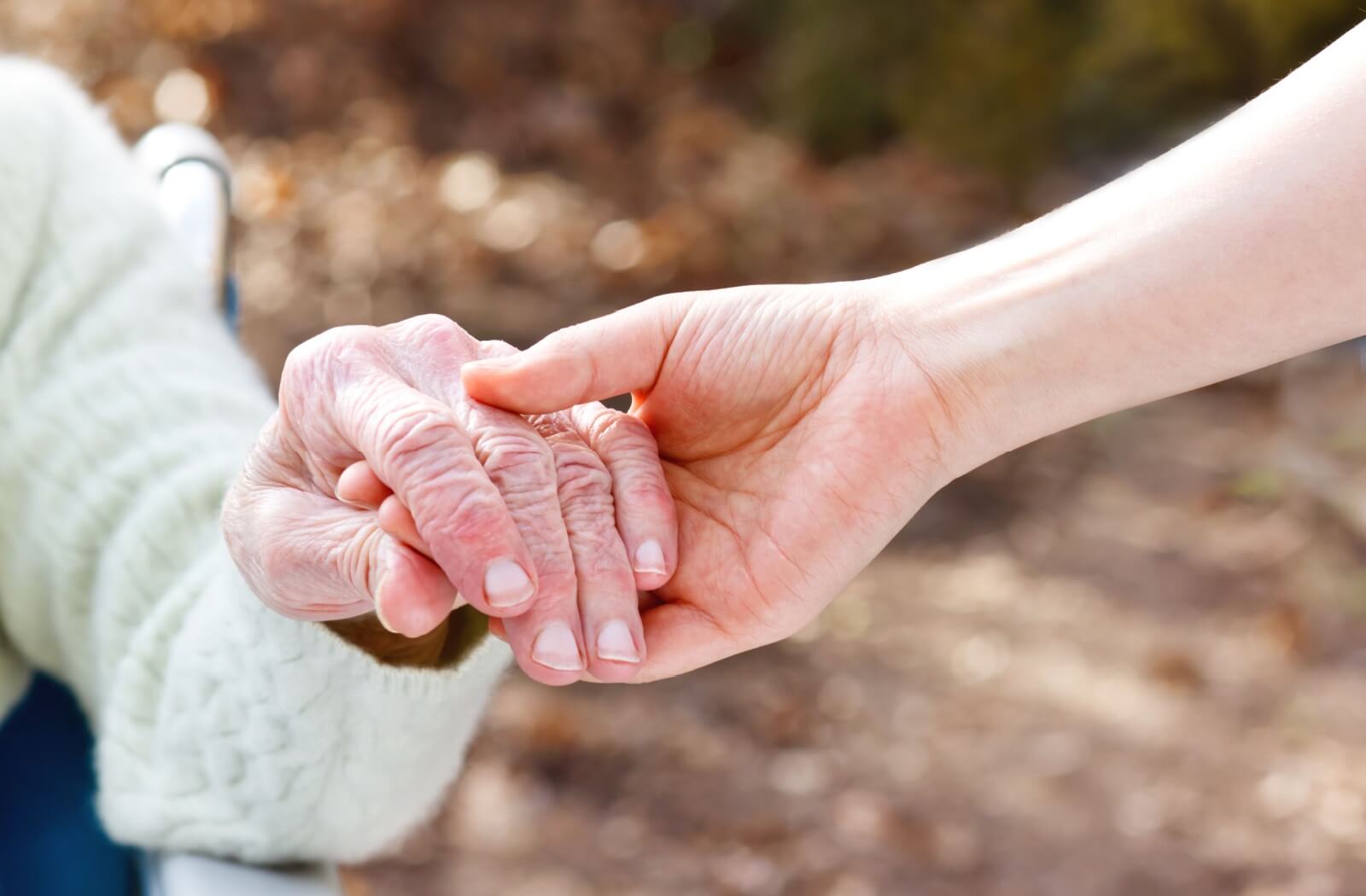 Close-up of a caregiver's hand gently holding an older adult's hand outdoors, symbolizing compassion and memory care support