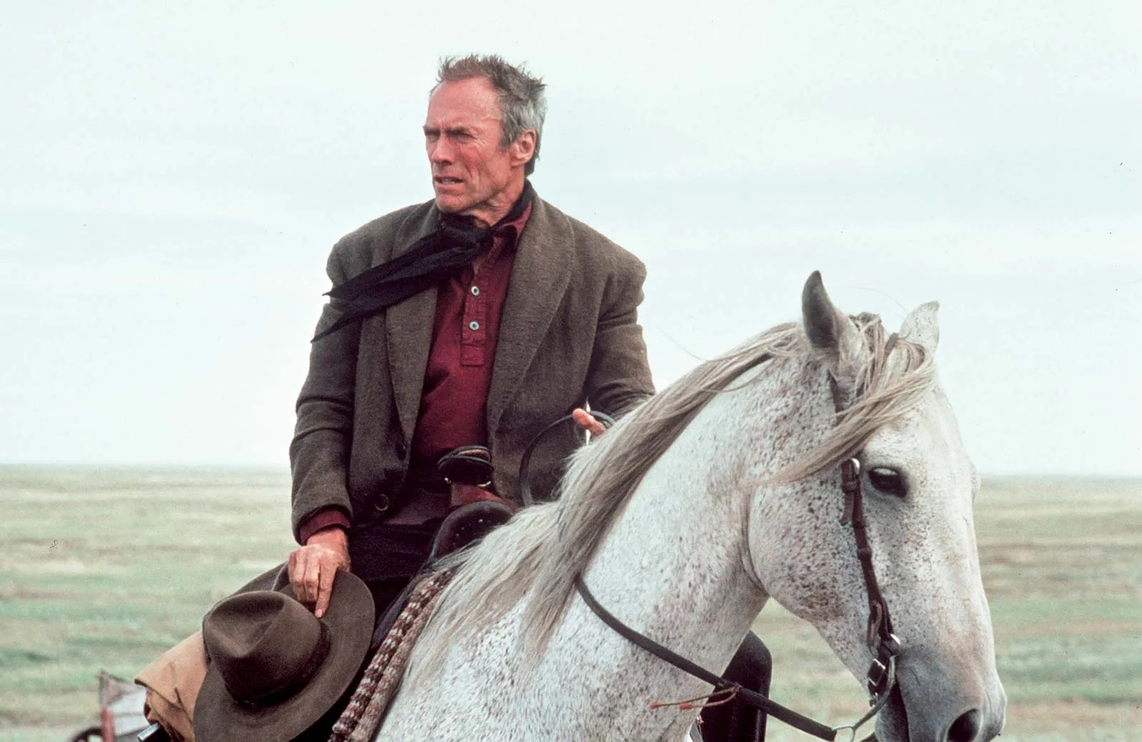 Clint Eastwood montando un caballo blanco en un paisaje rural, vestido con ropa de época de vaquero, capturado en una escena de una película del oeste.