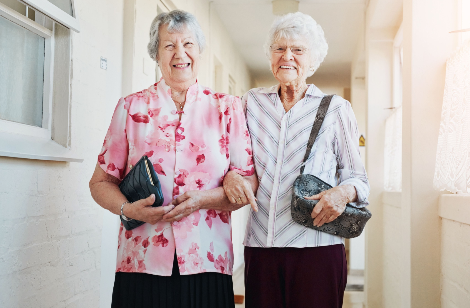 2 seniors stand arm-in-arm in summery blouses, smiling bright.