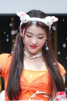 This contain Xiaoting  with long brown hair wearing an orange shirt and bunny ears on her head