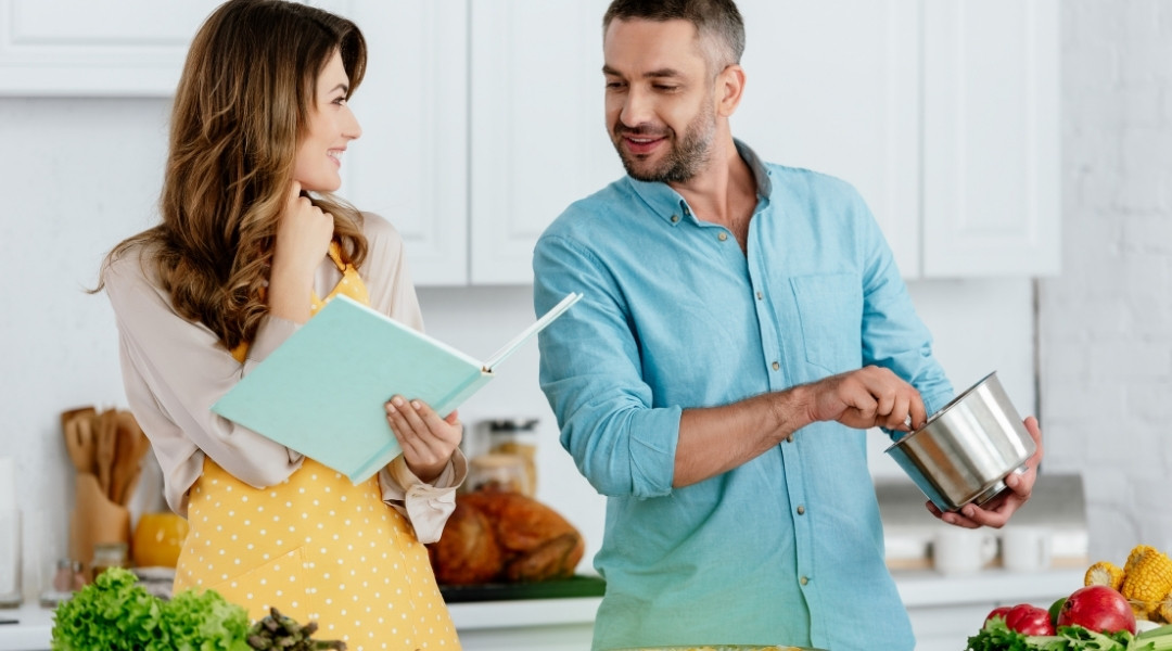 Un couple souriant cuisine ensemble dans une cuisine lumineuse; la femme tient un livre de recettes et l'homme mélange dans une casserole.