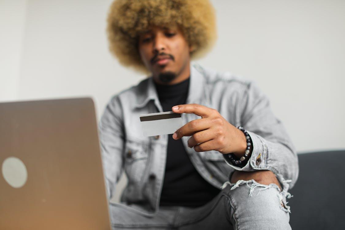 Free A man with afro hair using a credit card for online shopping while sitting indoors with a laptop. Stock Photo