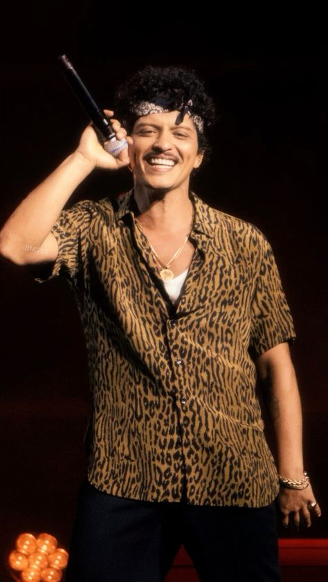 This contains a picture of Bruno Mars in a leopard print shirt holding a mic to his ear and smiling