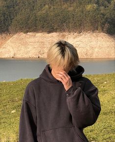 This contains an image of Kim Tae I  standing in front of a body of water wearing a black sweatshirt and holding their hand to his face