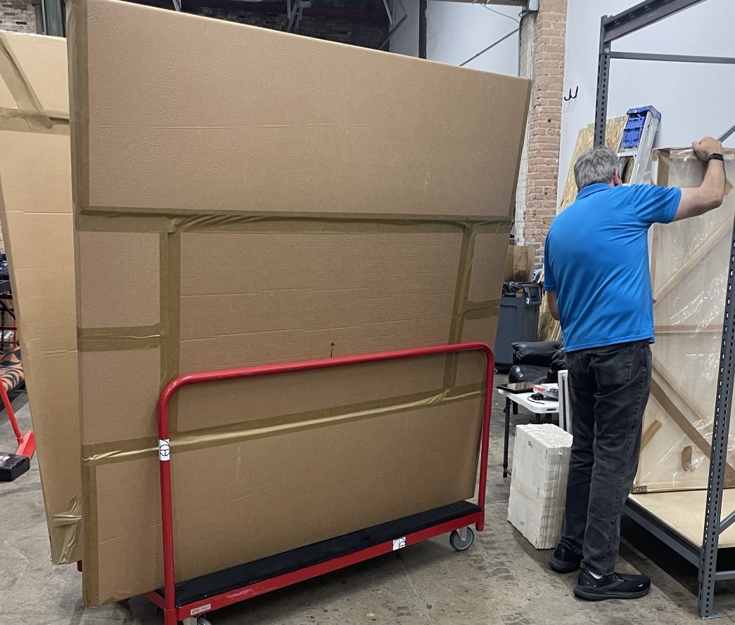 A man in a blue shirt beside a big brown package on a red trolley.