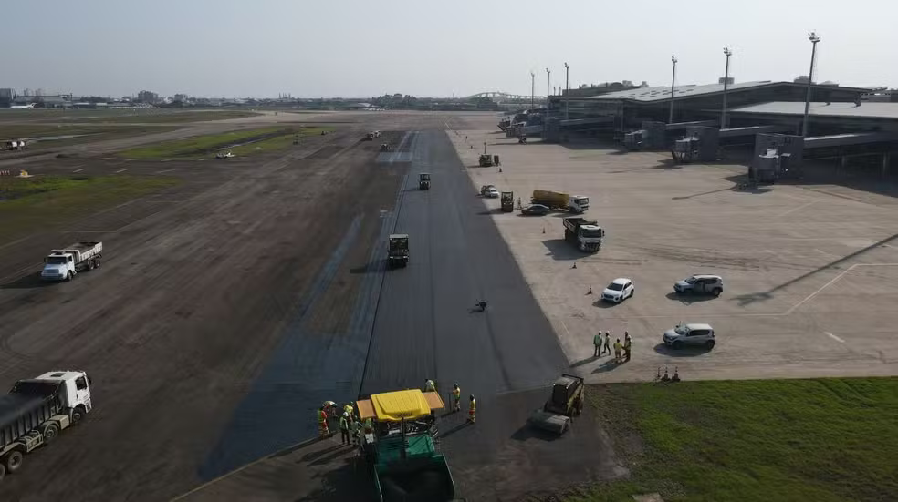 Obras de recuperação da pista do Aeroporto de Porto Alegre, com trabalhadores e máquinas pavimentando trecho de asfalto, preparando para retomada das operações comerciais.