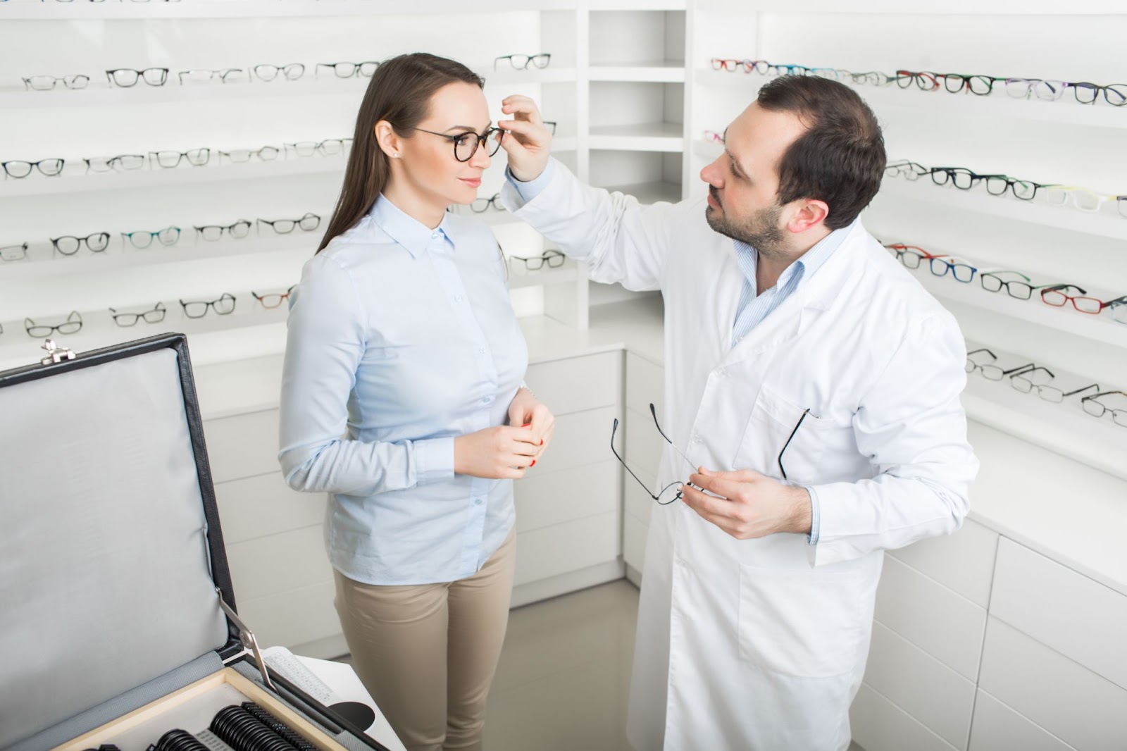 Woman chooses eyeglasses to correct vision standing at ophthalmological store near shelf with many glasses. Doctor ophthalmologist helps the patient to choose a frame for glasses.