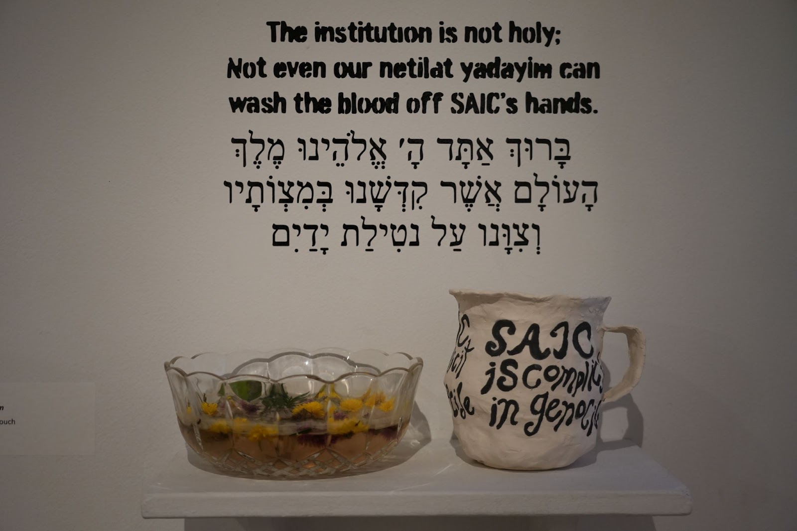 Image: Black text on a white wall reads, “The institution is not holy; Not even our netilat yadayim [Jewish ritual handwashing] can wash the blood off SAIC’s hands.” On a table in front of the text sits a glass bowl with water and flowers in it and a white jug with black writing stating, “SAIC is complicit in genocide.” Photo by Curators Under Censorship. 