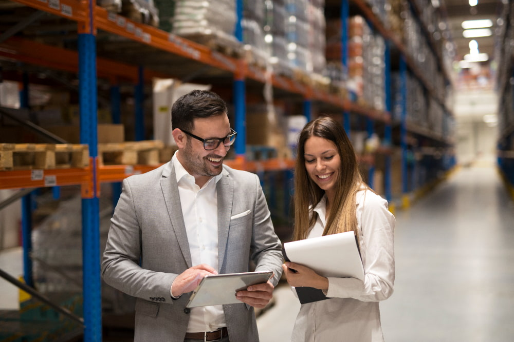 A man and woman with a clipboard smiling and conversing in a clearly organized warehouse filled with shelves of boxes.