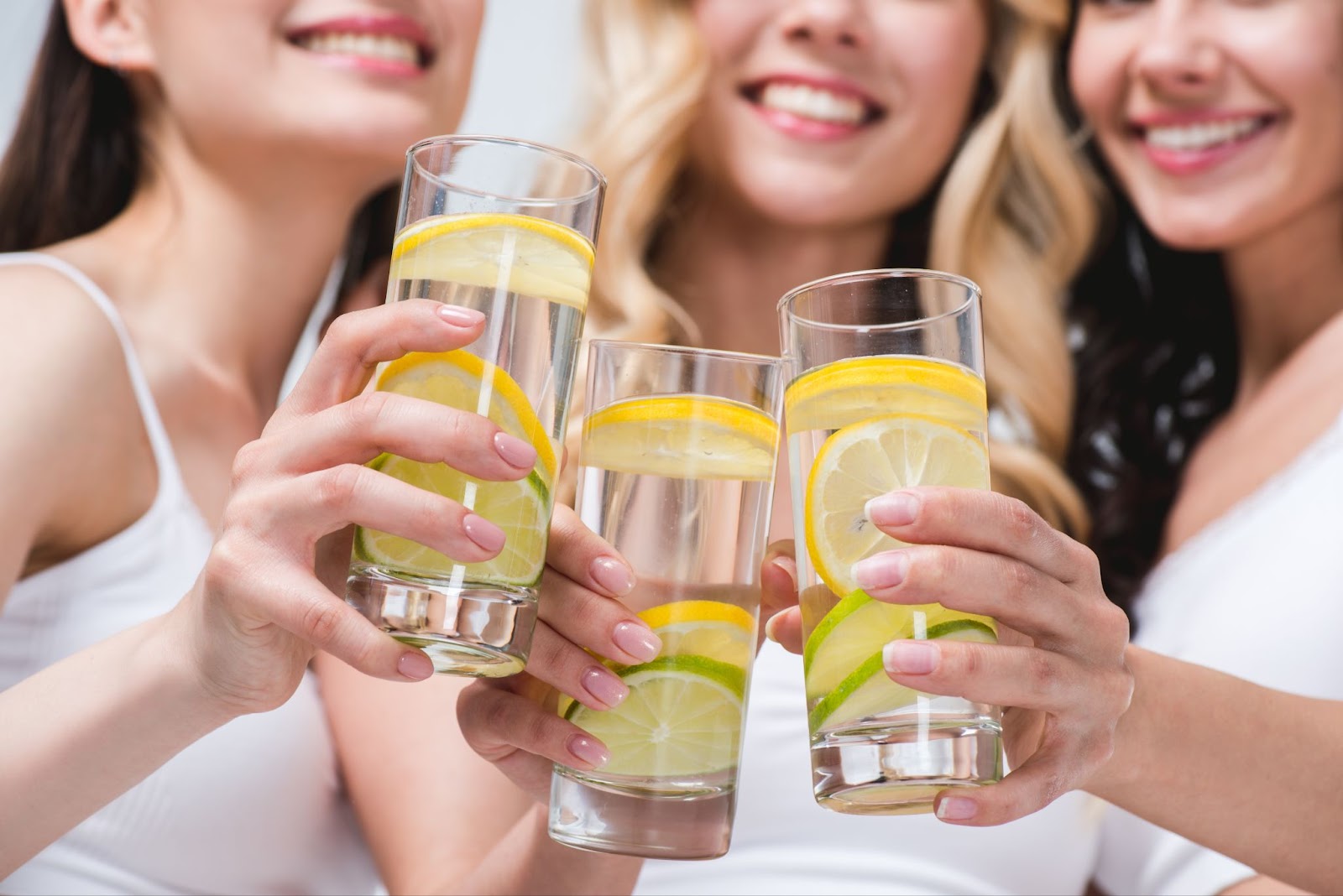 Three smiling young women clinking glasses of lemon water together.