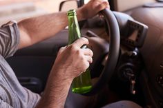 This contains an image of a man holding a green bottle while driving a car