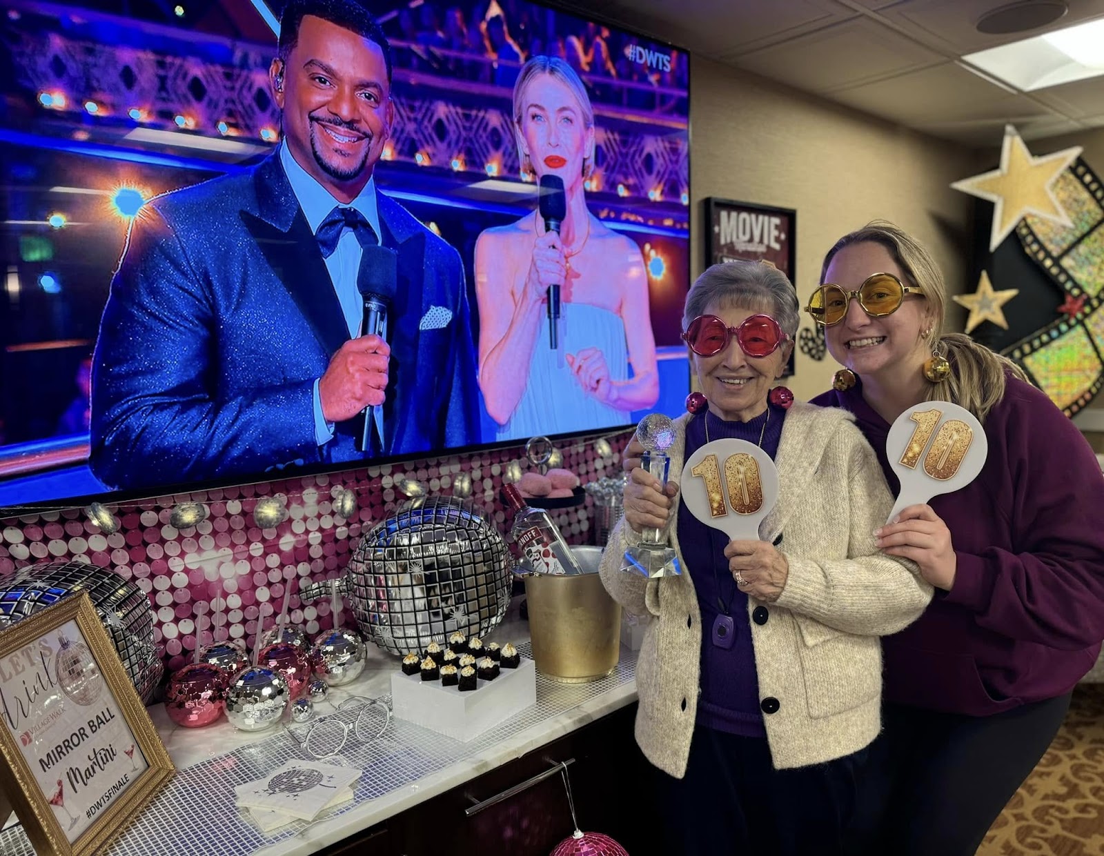 A picture of an assisted living caregiver holding an award with a resident in front of a TV