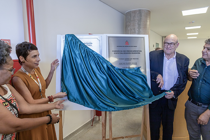 A imagem mostra um evento de inauguração, possivelmente da Farmácia de Medicamentos Especializados Diadema. Um grupo de quatro pessoas está participando da cerimônia, revelando uma placa comemorativa coberta por um tecido azul brilhante.  A placa, ao ser descerrada, exibe informações relacionadas ao Governo do Estado de São Paulo e menciona nomes como "Tarcísio Gomes de Freitas" e "José de Felipe Junior", indicando autoridades envolvidas na inauguração ou gestão do local. O ambiente é formal e bem iluminado, com paredes claras e um corredor ao fundo.  As pessoas presentes estão vestidas de forma apropriada para o evento, com roupas formais ou profissionais. Dois dos participantes seguram o tecido azul enquanto o retiram da placa, simbolizando a inauguração oficial do local. O clima parece ser de celebração e reconhecimento pela entrega do serviço à comunidade.