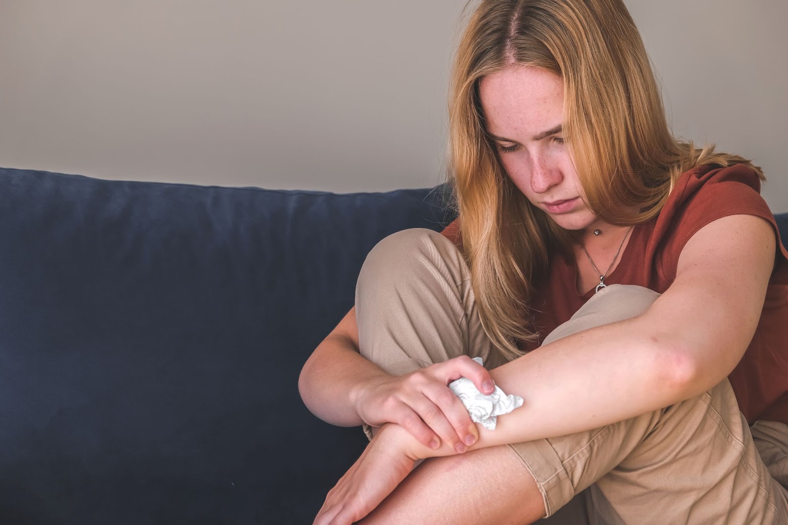 A woman sits on a couch, hugging her knees and holding a tissue, gazing downward with a look of depression or frustration.