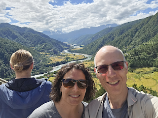 Man and woman smiling at the camera. In the distance behind them is a river running through a valley between mountains