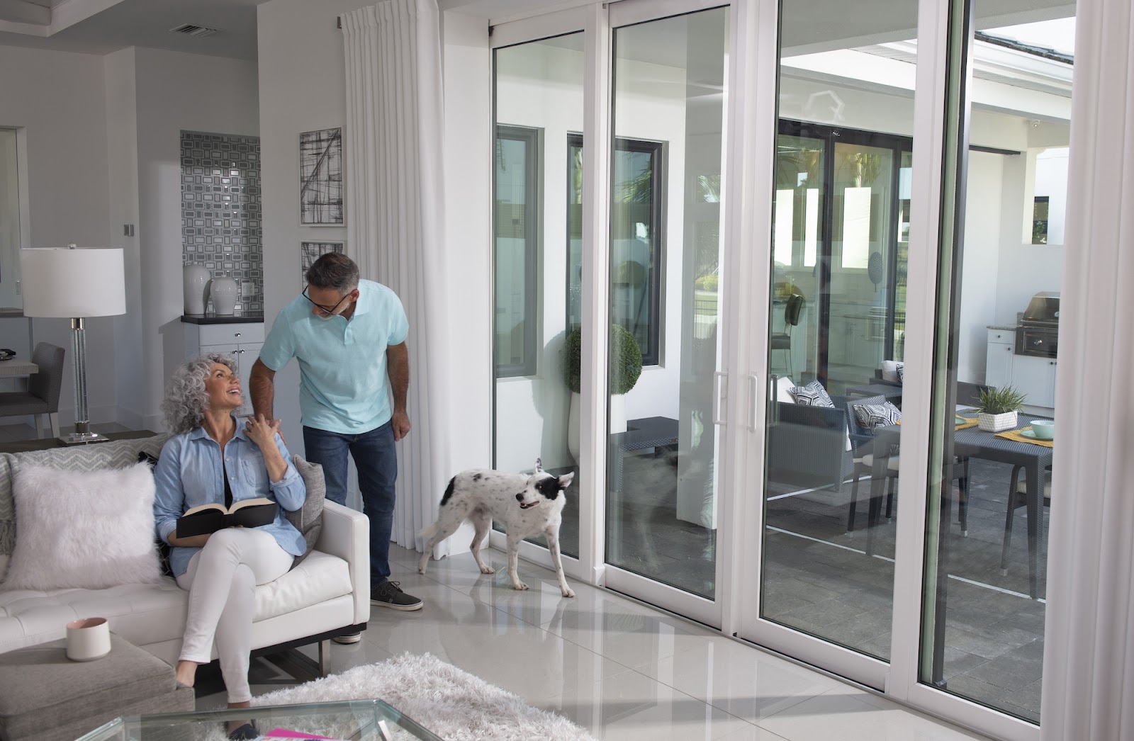 Two people and a dog relax inside their house. A set of closed exterior sliding glass doors separates them from their patio.