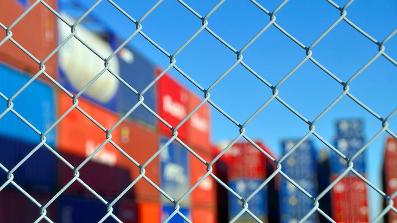 A sharp chain-link fence in front, with blurred colorful shipping containers and a clear blue sky in the background.