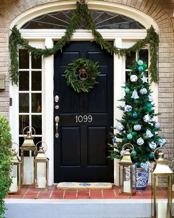 brick house with black door and a Christmas wreath a swag of greens and a christmas tree and lanterns