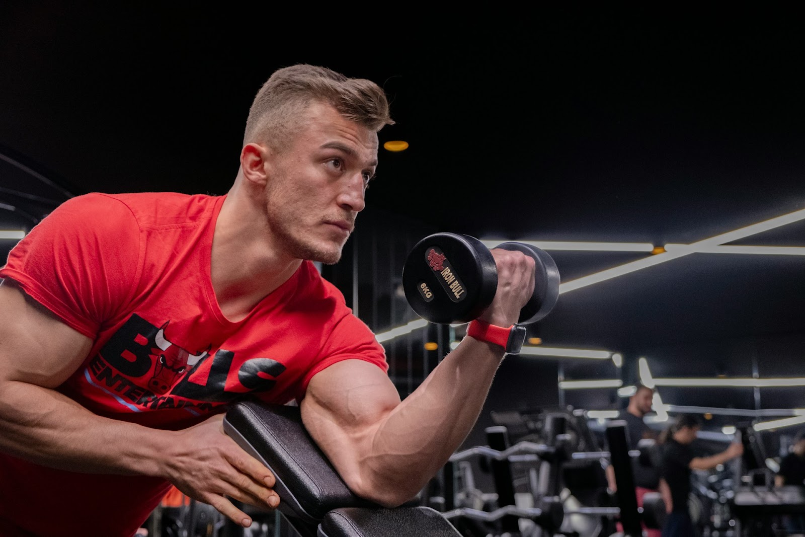 A  man wearing a red t-shirt in a gym holding a dumbbell.