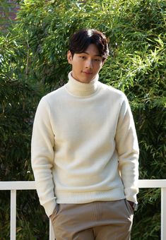 This contain a picture of Kim Ji Soo in a white turtle neck sweater and khaki pants standing on a porch