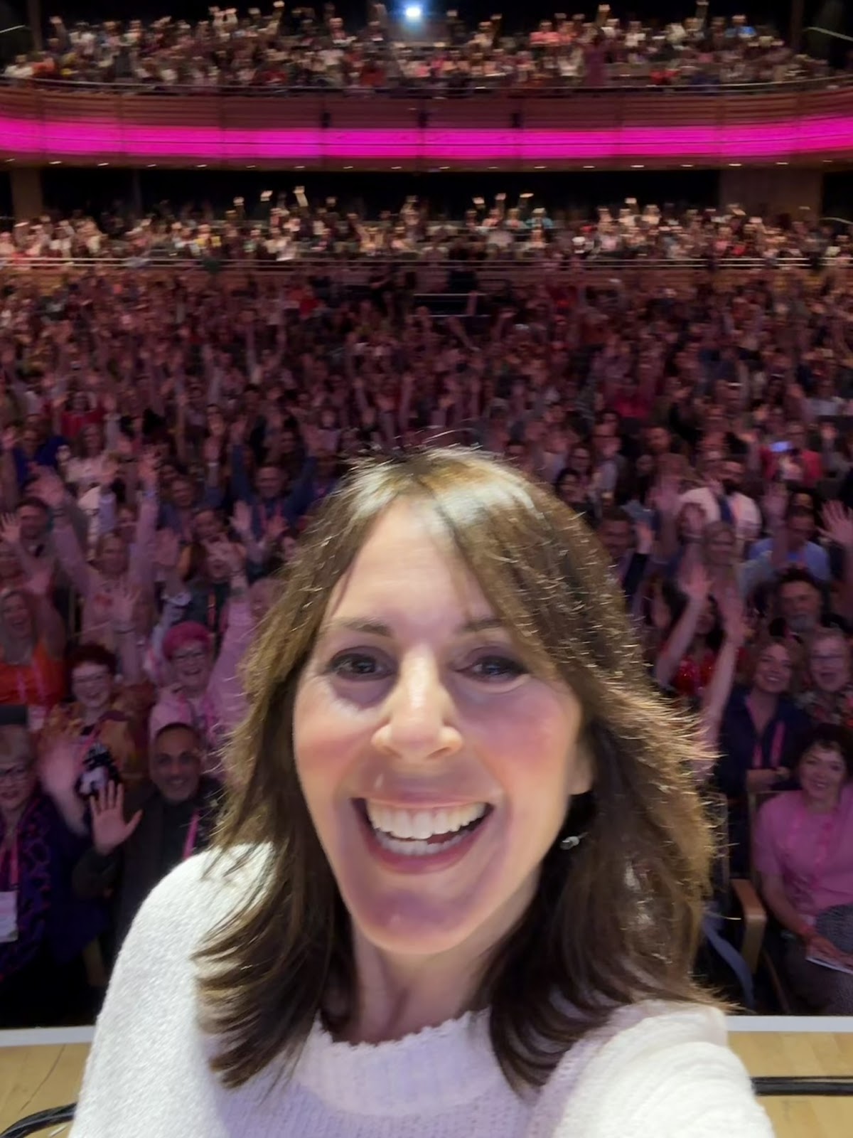 Laura Belgray takes a smiling selfie from a stage, she has on a white long-sleeve sweater with shoulder length brown hard. In the selfie background is a large, enthusiastic audience in a brightly lit auditorium. The crowd behind her is packed and animated, many waving and smiling toward the camera. The upper balcony is also filled with attendees, and pink lighting accents the ceiling and railings, creating a vibrant and celebratory atmosphere.