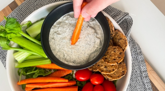 A hand dipping a carrot stick into a black bowl of Greek yogurt ranch dip, surrounded by celery sticks, cherry tomatoes, and whole-grain crackers.