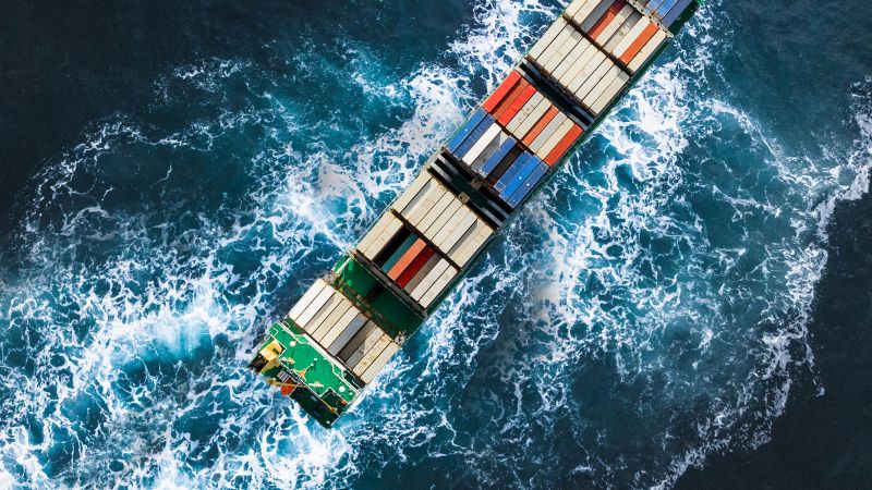 Aerial view of a cargo ship with colorful containers, creating white waves as it moves through the ocean.