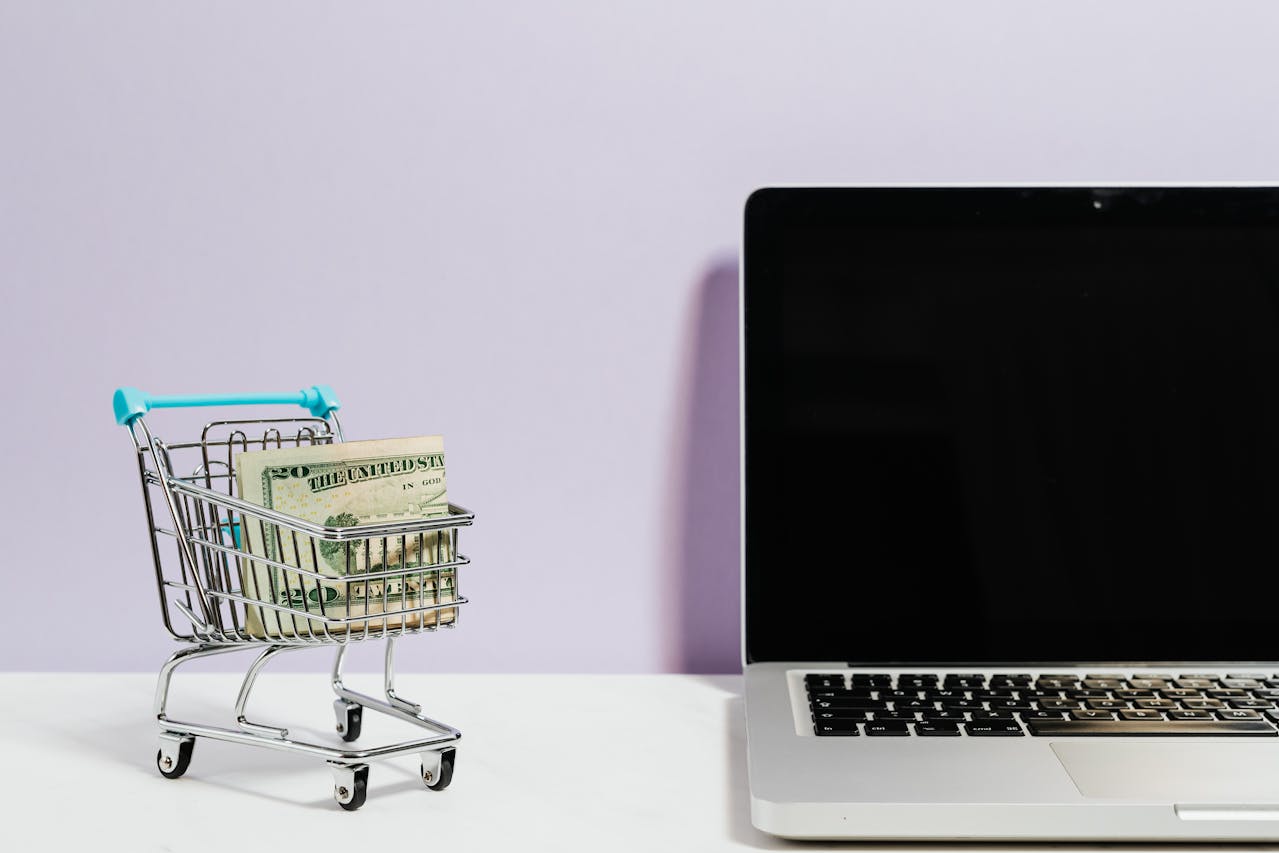 A mini shopping cart filled with US dollar bills placed beside a laptop, representing ecommerce business growth.