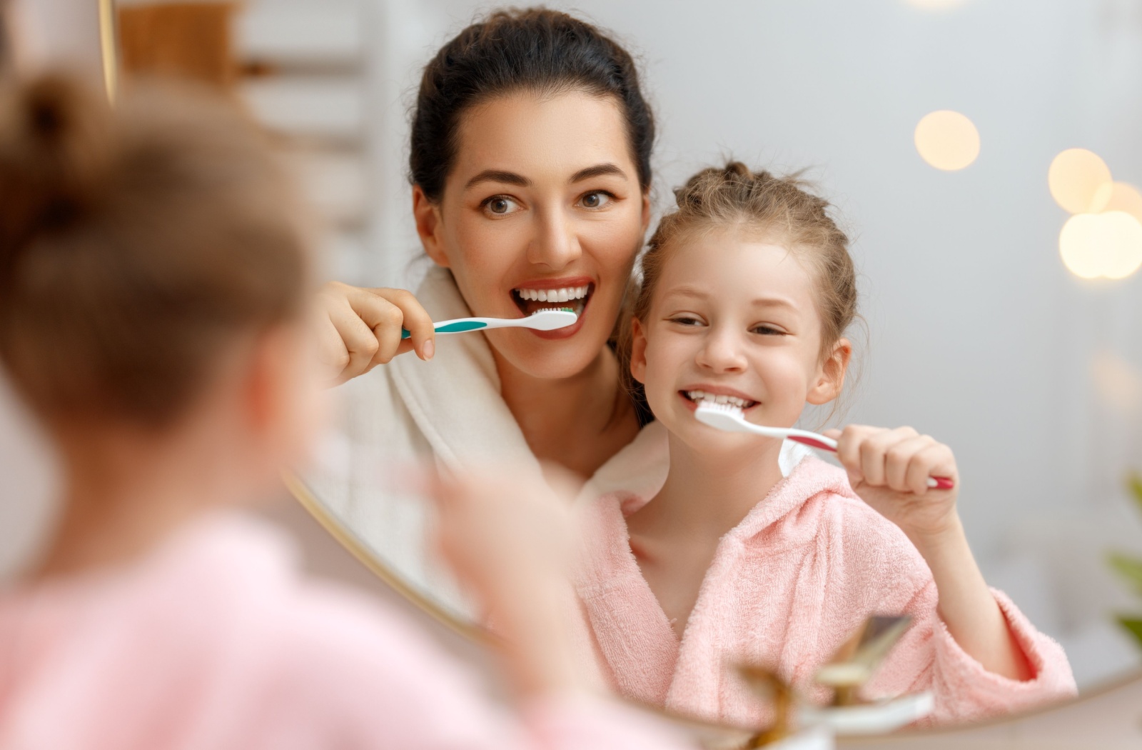 A parent and child smiling as they brush their teeth together in front of a bathroom mirror.