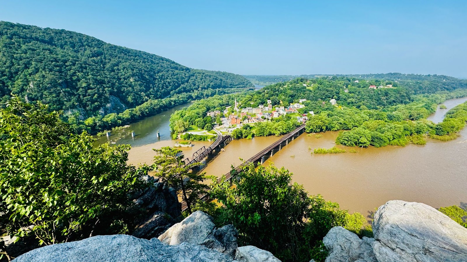 View from a rocky overlook with two rivers, one muddy and one clear, below. There is a small town at the center of the image, a mountain to the left, and clear skies overhead.
