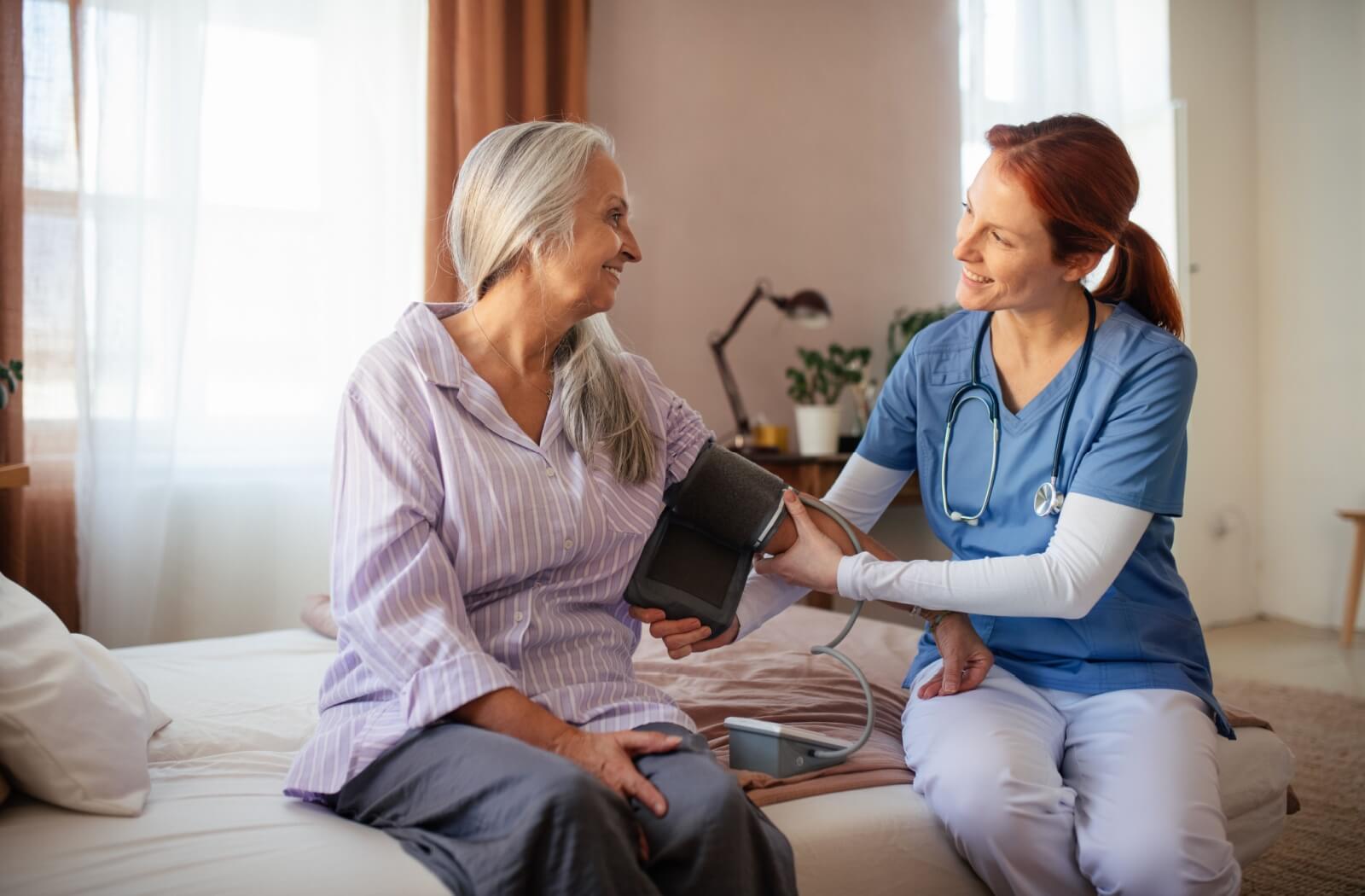 A patient holds out their arm for their nurse to take their blood pressure while both sit on the patient’s bed at home