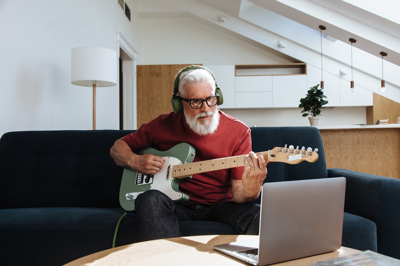 A man playing an electric guitar while wearing headphones, using his laptop to create AI music in a modern, well-lit living room.