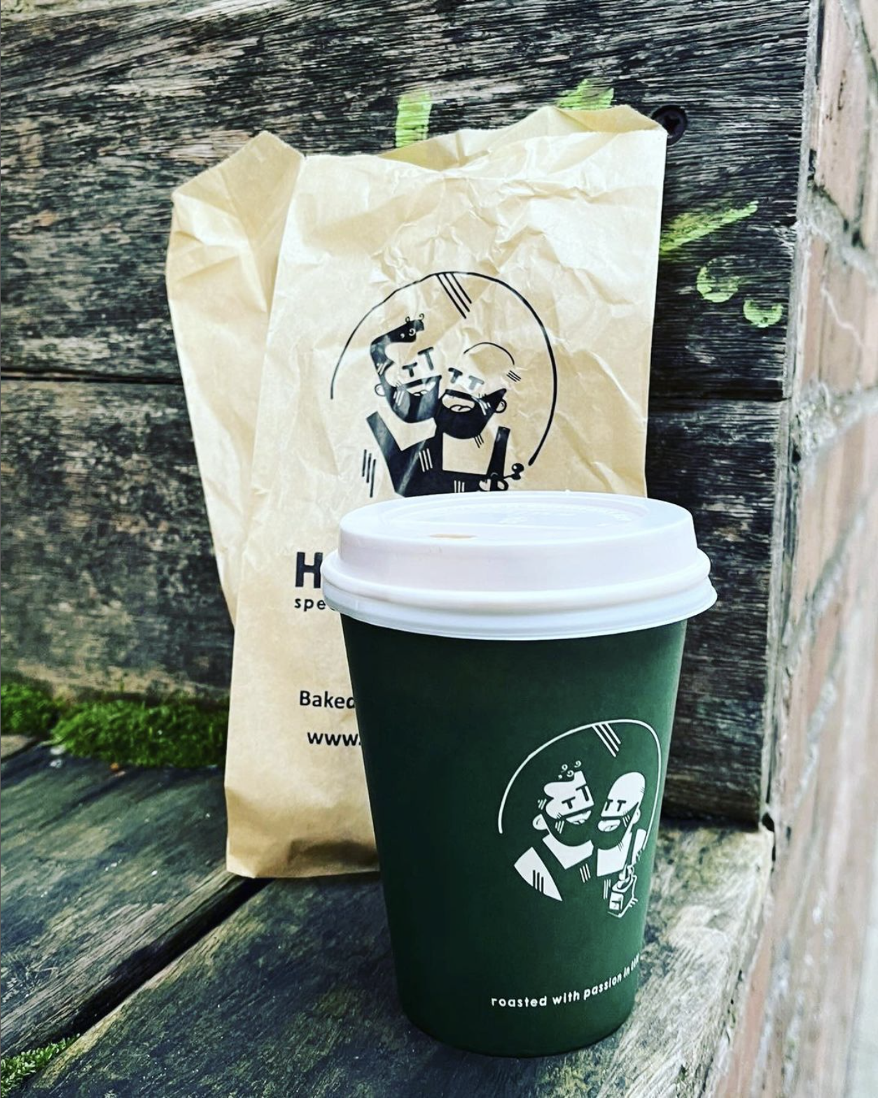 A green coffee cup with a white lid, featuring a minimalist illustration of two bearded men, set on a rustic wooden surface with a brown paper bag in the background.