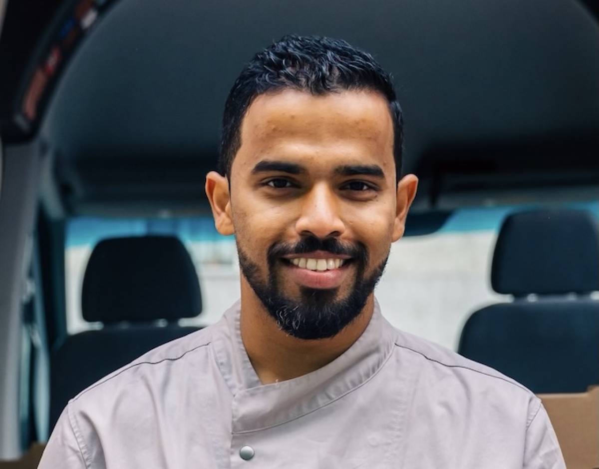 Chef Vish Mayekar smiles at the camera, wearing chef whites, with car head rests behind him