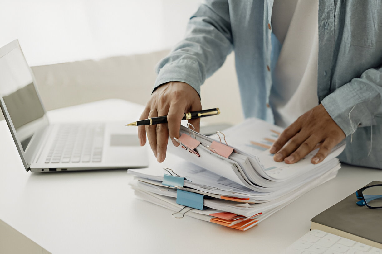 Close-up of a person reviewing financial documents with a pen in hand next to a laptop, representing bookkeeping organization for tax preparation.