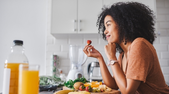 A smiling woman eating fresh fruit in a modern kitchen, surrounded by orange juice and other healthy foods.