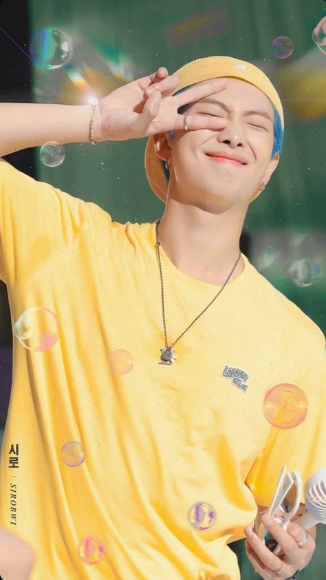 This  contain BTS RM  wearing a yellow shirt and headband holding a hair dryer in front of his face