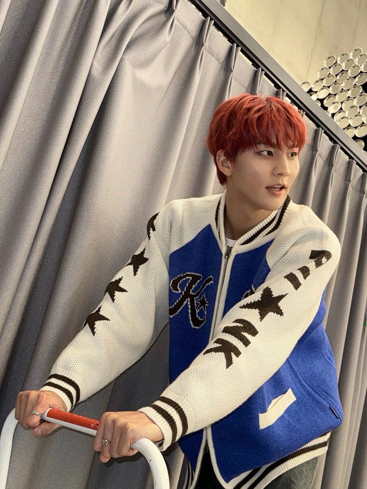 This contains an image of VANNER Hyesung's in a blue and white jacket is holding a red handlebar while standing next to a curtain