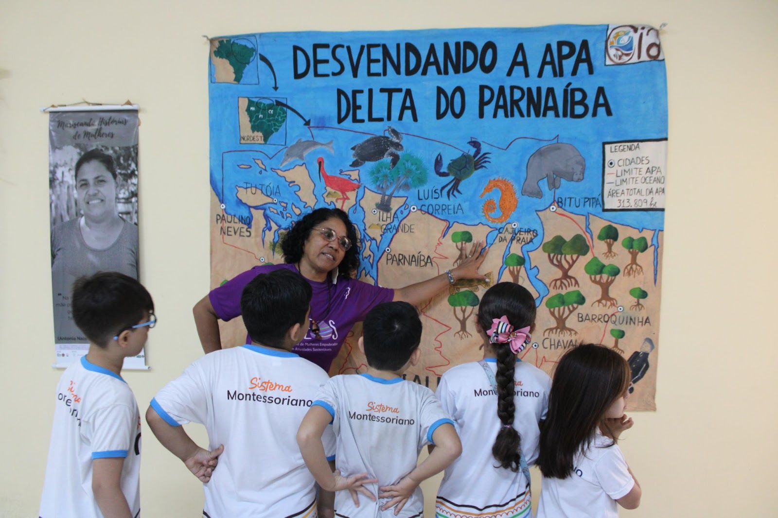 Em uma parede branca, há cartazes e banners da exposição 
"Mariscando Histórias e Mulheres", sobre marisqueiras. Uma mulher está apontando a mão para um dos cartazes, em frente a estudantes.
