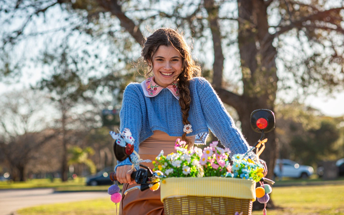 Joven sonriente con suéter azul montando bicicleta con una canasta de flores, disfrutando de un día soleado en un parque.