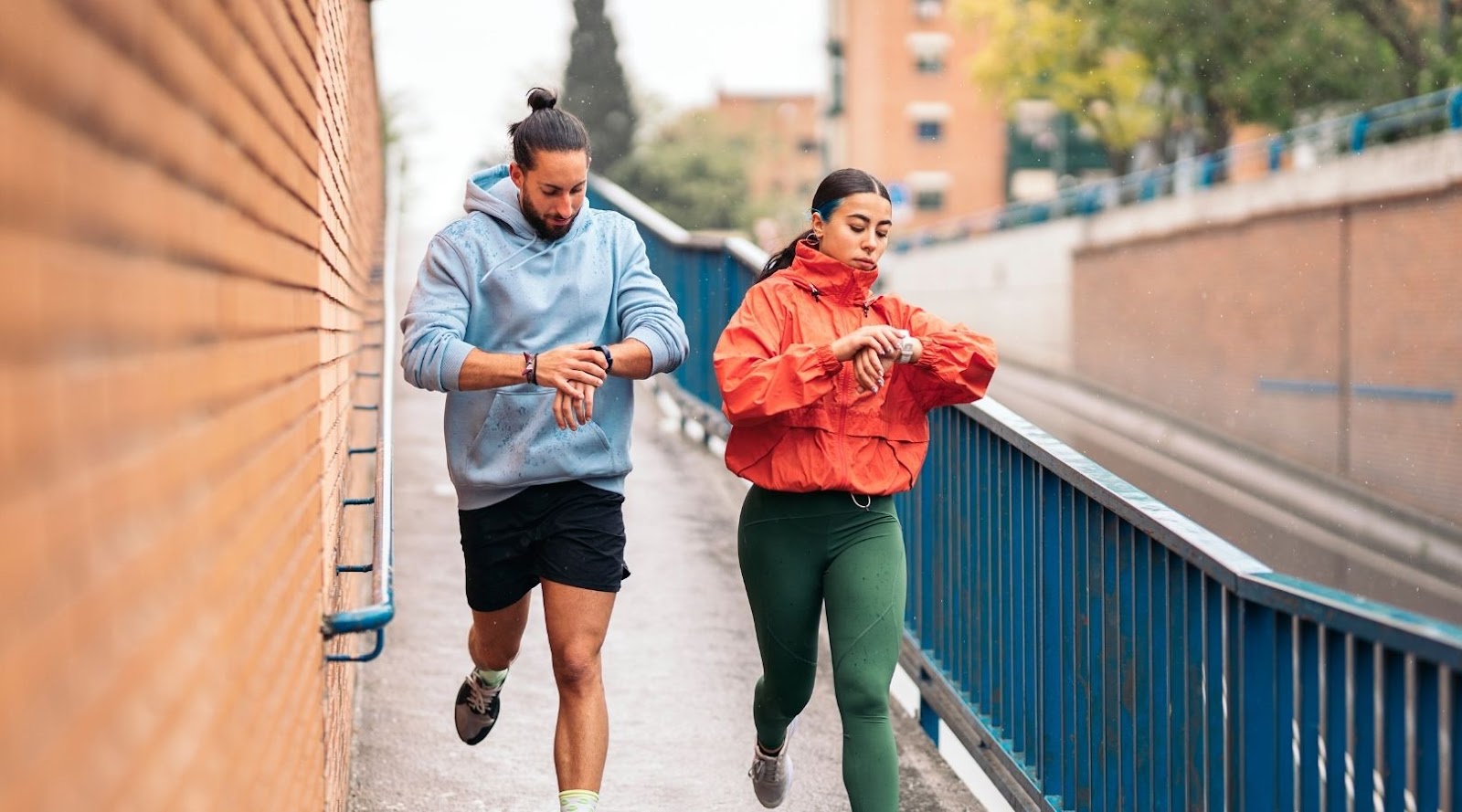 Deux coureurs vérifiant leurs montres intelligentes tout en joggant sous la pluie, vêtus de chandails à capuche sportifs, sur un sentier urbain.